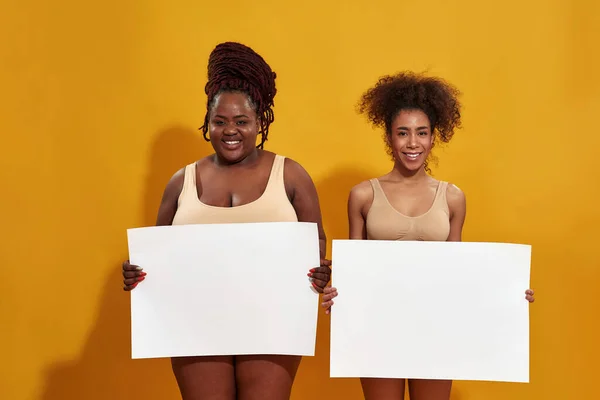 Dos jóvenes mujeres afroamericanas hermosas en ropa interior sonriendo a la cámara, sosteniendo una pancarta en blanco para su texto, de pie aislada sobre un fondo amarillo — Foto de Stock