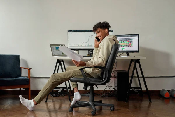 Ganzkörperaufnahme eines entspannten jungen männlichen Händlers, der Papiere mit Statistiken hält, während er telefoniert, am Schreibtisch sitzt und von zu Hause aus handelt — Stockfoto
