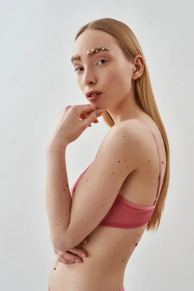 Foto creativa de modelo femenino caucásico joven natural con flores en las cejas con sujetador transparente rosa mirando a la cámara, posando aislado sobre fondo gris claro —  Fotos de Stock