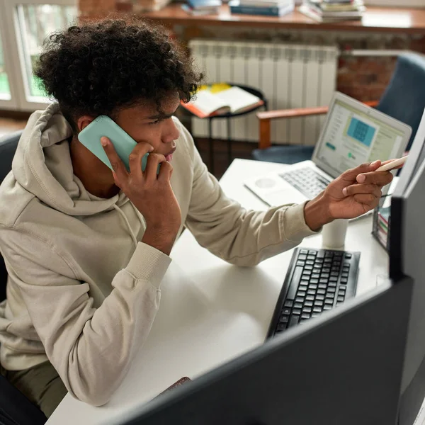 Hochwinkelaufnahme eines geschäftigen jungen männlichen Aktienhändlers, der mit dem Kunden telefoniert, während er den Computer benutzt, und zu Hause am Schreibtisch im Wohnzimmer sitzt — Stockfoto