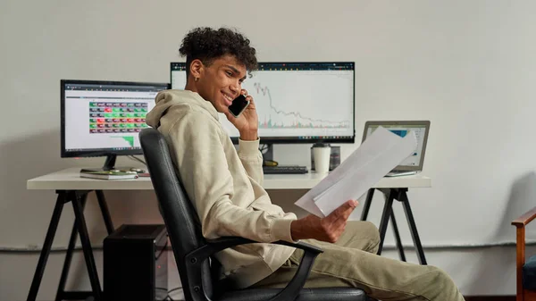 Erfolgreicher junger männlicher Händler, der Papiere hält, lächelnd mit einem Kunden telefoniert, am Schreibtisch sitzt und von zu Hause aus handelt — Stockfoto