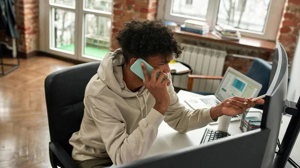 Hochwinkelaufnahme eines geschäftigen jungen männlichen Aktienhändlers, der mit dem Kunden telefoniert, während er den Computer benutzt, und zu Hause am Schreibtisch im Wohnzimmer sitzt — Stockfoto