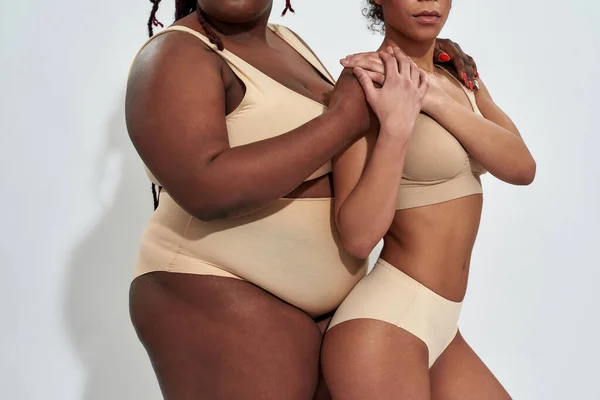Foto recortada de cuerpos de dos mujeres afroamericanas en ropa interior beige con diferente peso apoyándose mutuamente, posando juntas aisladas sobre fondo gris —  Fotos de Stock