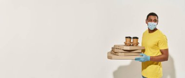 Koruyucu maske takan erkek kurye ve eldivenler kameraya bakıyor, pizza kutuları ve içecekler taşıyor, gri arka planda yalnız poz veriyor.