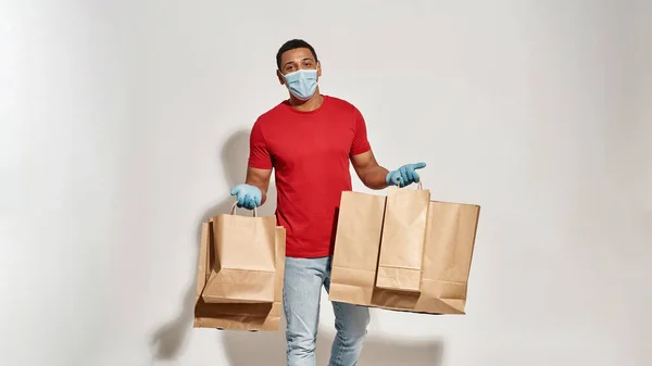 Wesoły dostawca w masce ochronnej i rękawiczkach, trzymający wiele brązowych papierowych toreb na wynos, stojący na jasnoszarym tle — Zdjęcie stockowe