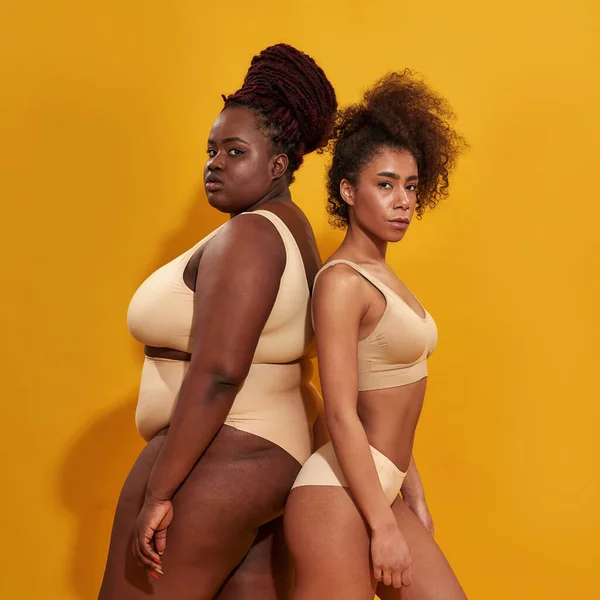 Två starka afrikanska amerikanska kvinnor i beige underkläder med olika kroppsstorlek står tillsammans rygg mot rygg, poserar isolerade över gul bakgrund — Stockfoto