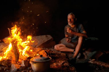 Vahşi doğada kamp ateşinin yanında oturmuş yemek yiyen aç bir adam.