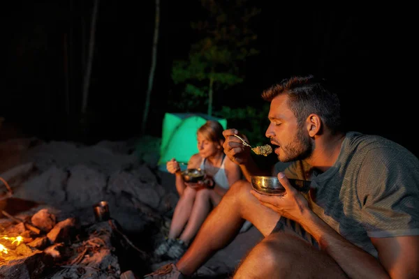 Retrato de turista cansado comendo uma refeição cozida no fogo enquanto relaxa junto com sua namorada na natureza — Fotografia de Stock