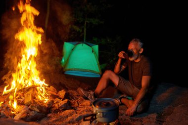 Olgun erkek turist sıcak çay ya da kahve içerken geceleri ormanda kamp ateşinin yanında dinleniyor.