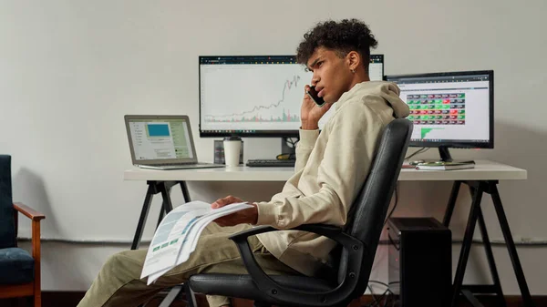 Seriöser junger männlicher Händler, der Papiere hält, während er mit einem Kunden telefoniert, am Schreibtisch sitzt und von zu Hause aus handelt — Stockfoto