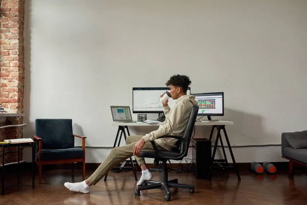 Largura completa de tiro relajado joven comerciante masculino beber café mientras está sentado en el escritorio, el comercio en línea desde casa — Foto de Stock
