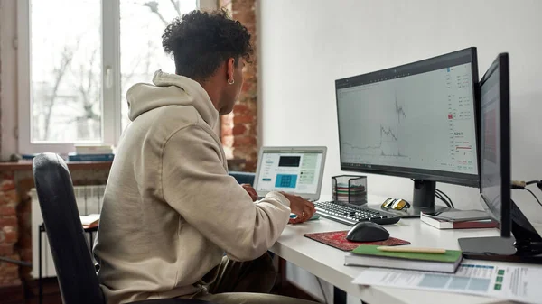 PCとラップトップを使用してオンラインで取引若い男の側面図,現代のリビングルームのオフィスに家に座って — ストック写真