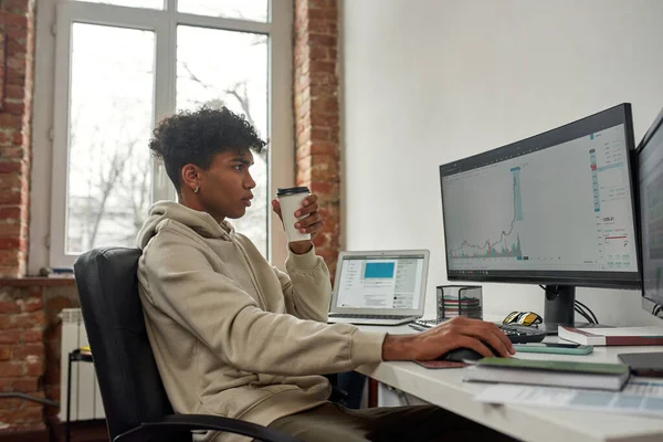 Vista laterale del giovane commerciante maschile che beve caffè, lavora al computer, studia grafici mentre commercia da casa — Foto Stock