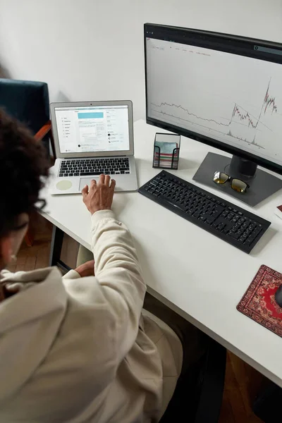 Vista trasera del comerciante masculino joven usando el ordenador portátil y la PC, mirando a la pantalla del ordenador para analizar las estadísticas mientras está sentado en el escritorio de la oficina en casa — Foto de Stock