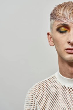 Parlak karanlık parti makyajlı genç transseksüelin yarı yüzü beyaz arka planda kapalı gözlerle poz veriyor.