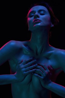 Çıplak, zayıf, göğsünü tutkuyla sıkan genç bir kadının portresi. Siyah arka planda mavi, neon bir ışıkla izole edilmiş şekilde poz veriyor.