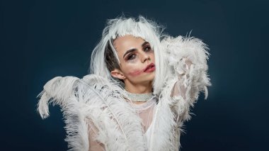 Yorgun, üzgün travesti gece makyajlı, boa tüyü parti elbisesi giyen, mavi arka planda soyutlanmış beyaz peruğu olan genç travesti.