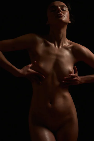 Apaixonado modelo nu feminino de pé com os olhos fechados, tocando seu peito enquanto posando isolado no fundo escuro — Fotografia de Stock