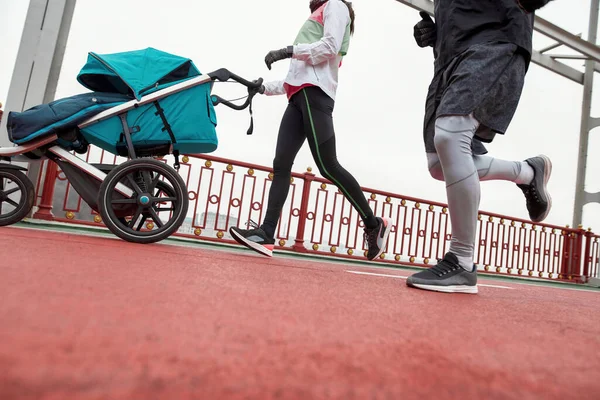 Tiro recortado de pessoas de esportes atléticos, pais jovens que treinam em conjunto com um carrinho de criança em uma ponte na cidade em um dia nublado — Fotografia de Stock