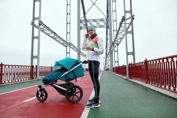 Filmagem completa de uma jovem motivada usando fones de ouvido enquanto estava na ponte com uma carruagem de bebê, preparada para correr em um dia nublado na cidade — Fotografia de Stock