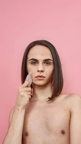 Bastante joven transexual con cabello castaño oscuro usando piedra de gua sha de cuarzo rosa para masaje facial, posando aislado sobre fondo rosa —  Fotos de Stock