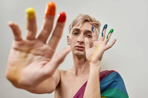 Joven transexual mirando a la cámara y posando con los dedos en colorido arco iris lgbtqi pintura aislada sobre fondo claro —  Fotos de Stock