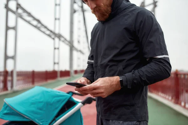 Tiro recortado de pai corredor masculino usando smartphone enquanto se prepara para uma corrida na cidade com carrinho em um dia nublado — Fotografia de Stock