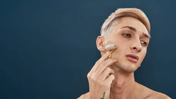 Porträt eines androgynen Transgender-jungen Mannes mit hellen Haaren, der eine Quarzwalze zur Gesichtsmassage benutzt und wegschaut, während er isoliert vor dunkelblauem Hintergrund posiert — Stockfoto