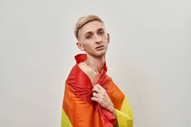 LGBT 'nin renkli gökkuşağı bayrağına sarılı genç transseksüelin portresi ışık arkaplanı üzerinde izole bir şekilde poz verirken kameraya bakıyor.