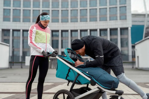 Cuidar de jovens pais esportivos colocando seu bebê em carrinho, andando juntos na cidade em um dia frio de outono — Fotografia de Stock