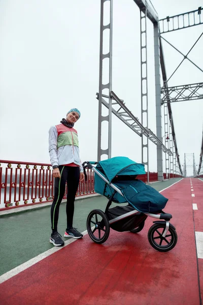 Foto completa de mujer joven calentándose, de pie en el puente con un carro de bebé, preparándose para trotar en un día nublado en la ciudad — Foto de Stock