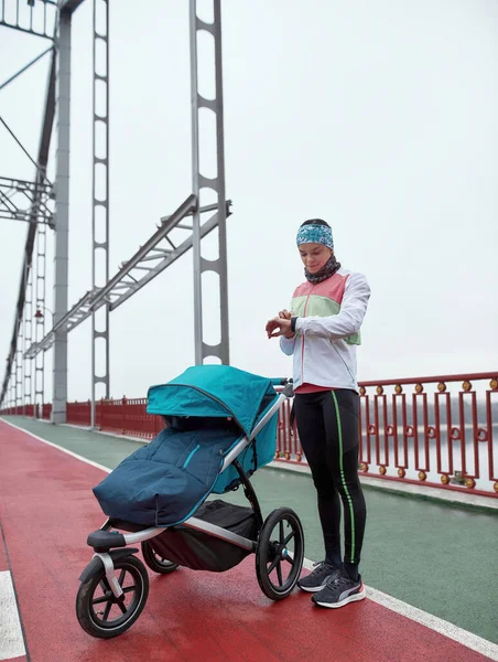 Tiro de comprimento total da jovem mulher verificando o tempo e os resultados usando relógio inteligente em seu pulso enquanto estava na ponte com uma carruagem de bebê em um dia nublado na cidade — Fotografia de Stock