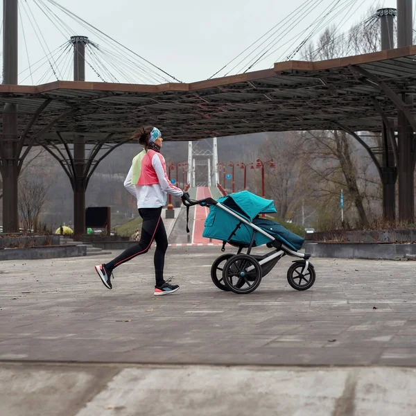Largura completa de tiro deportivo mamá corriendo y llevando un cochecito con un niño mientras se entrena en el parque en un frío día de otoño — Foto de Stock