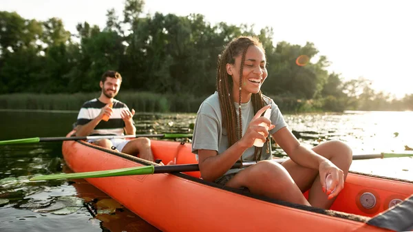 Vrolijke jonge vrouw met zonnebrandcrème fles, het aanbrengen van zonwering tijdens het kajakken op de rivier met haar vriendje buiten — Stockfoto