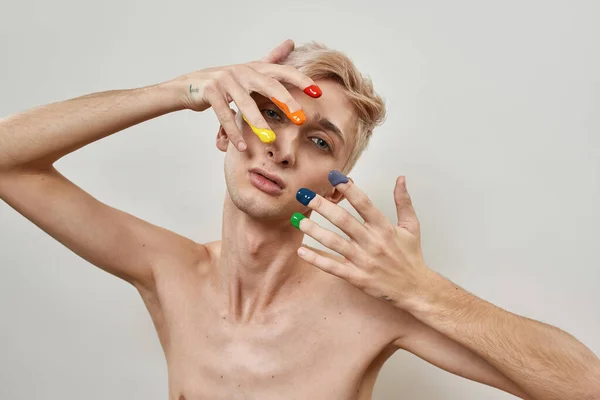 Retrato de joven transgénero con dedos en colorido arco iris lgbtqi pintura mirando a la cámara mientras posando aislado sobre fondo de luz —  Fotos de Stock