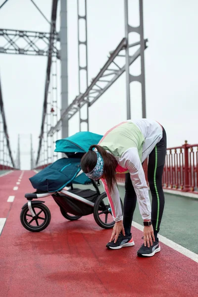 Tiro de comprimento total de esportista jovem ativo esticando seu corpo, de pé na ponte com uma carruagem de bebê, preparando-se para uma corrida em um dia nublado na cidade — Fotografia de Stock
