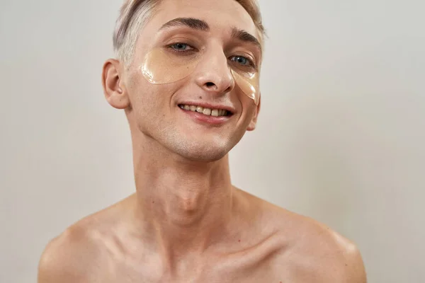 Portret szczęśliwego transseksualisty bez koszuli, młodego mężczyzny z nałożonymi pod oczy żelowymi łatkami, uśmiechniętymi do kamery i pozującymi na jasnym tle. — Zdjęcie stockowe