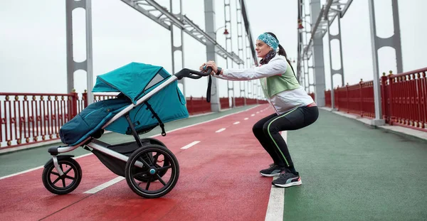 Tiro de comprimento total de esportiva jovem mãe esticando as pernas, exercitando-se na ponte com uma carruagem de bebê, preparando-se para uma corrida em um dia nublado na cidade — Fotografia de Stock