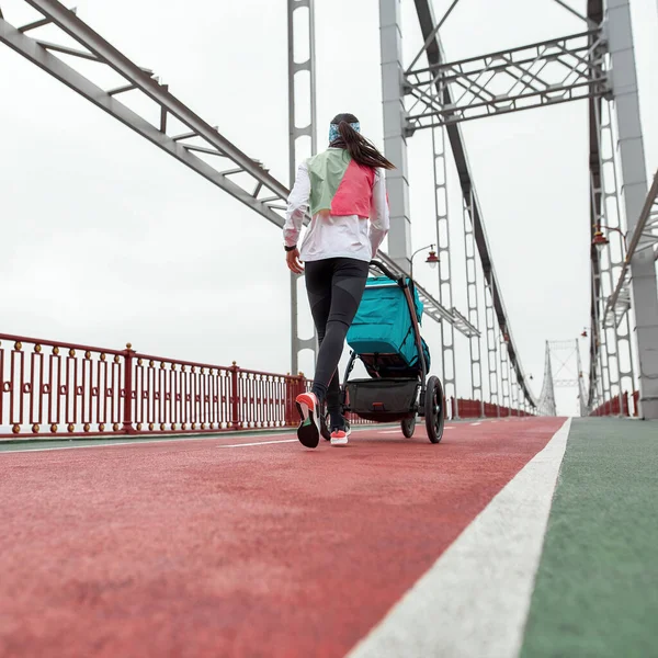 Mulher esportiva mãe correndo com uma carruagem de bebê em uma ponte na cidade em um dia nublado — Fotografia de Stock