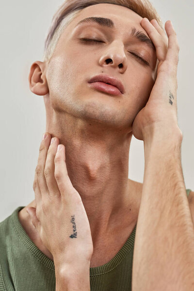 Закройте портрет чувственного андрогинного трансгендерного молодого человека с макияжем, трогающим его лицо, позируя с закрытыми глазами, изолированными на светлом фоне