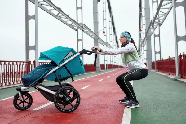Desportivo jovem mãe esticando as pernas, exercitando-se na ponte com uma carruagem de bebê, preparando-se para uma corrida em um dia nublado na cidade — Fotografia de Stock