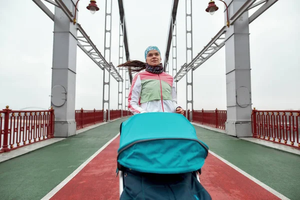 Motive olmuş genç sporcu anne şehirde bir köprüde antrenman yaparken bebek arabasıyla koşuyor. — Stok fotoğraf
