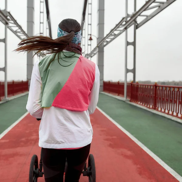 Visão traseira da jovem mãe ativa em sportswear correndo com um carrinho de criança enquanto treinava em uma ponte na cidade — Fotografia de Stock