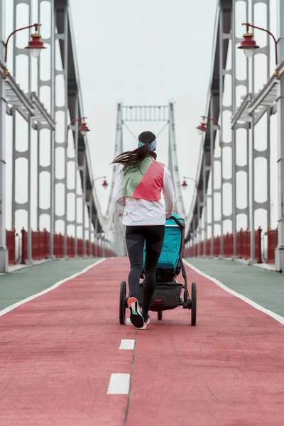 Visão traseira da mãe esportista correndo com um carrinho em uma ponte na cidade em um dia nublado — Fotografia de Stock