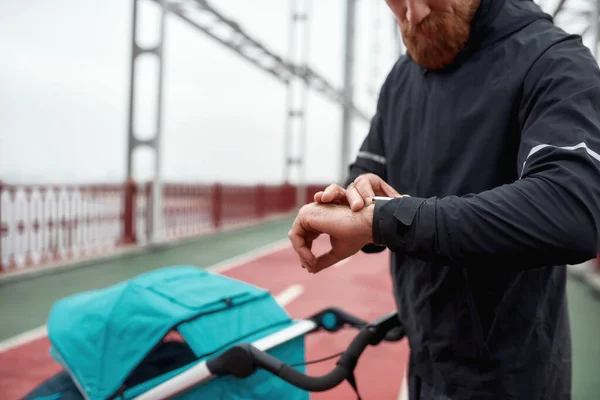 Tiro recortado de esportista verificando o tempo usando relógio inteligente em seu pulso enquanto caminhava na cidade com carruagem de bebê em um dia nublado — Fotografia de Stock