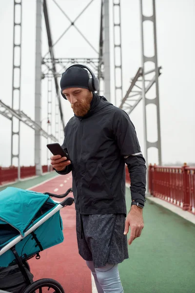 Pai esportivo usando fones de ouvido, usando smartphone enquanto se prepara para uma corrida na cidade com carrinho em um dia nublado — Fotografia de Stock