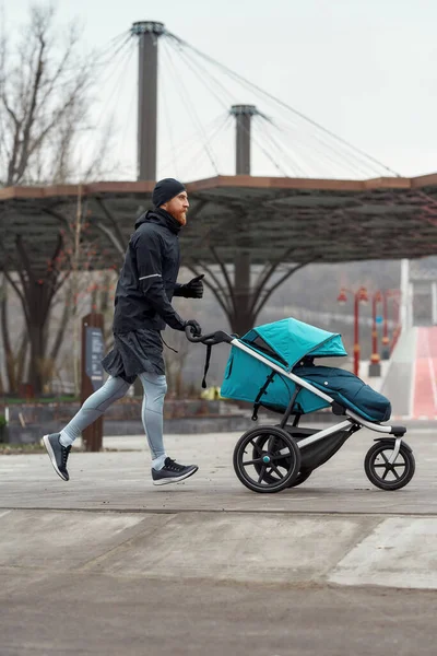 寒い秋の日に公園で訓練しながら、やる気のあるスポーティーなお父さんが走り、子供と一緒にベビーカーを運ぶフル長さのショット — ストック写真