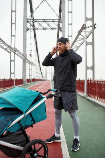 Tiro de comprimento total de pai jovem usando fones de ouvido enquanto estava na ponte com uma carruagem de bebê, pronto para uma corrida em um dia nublado na cidade — Fotografia de Stock