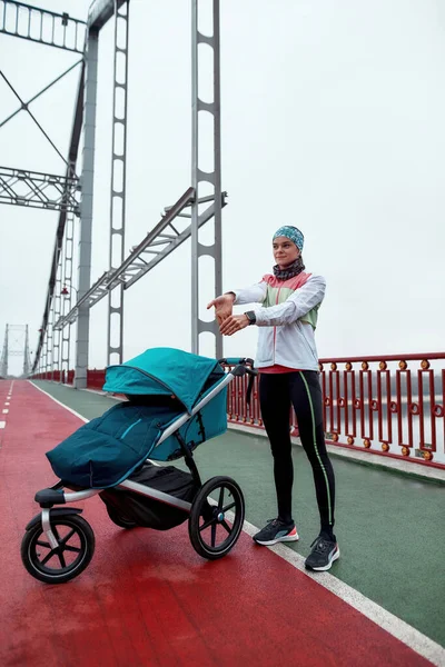 Tiro de comprimento total de jovem motivada esticando as mãos, de pé na ponte com uma carruagem de bebê em um dia nublado na cidade — Fotografia de Stock