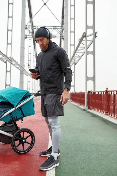 Πλήρες πλάνο του αθλητικού άνδρα, νεαρός πατέρας φορώντας ακουστικά, χρησιμοποιώντας smartphone ενώ ετοιμάζεται για τρέξιμο στην πόλη με καροτσάκι σε μια συννεφιασμένη μέρα — Φωτογραφία Αρχείου